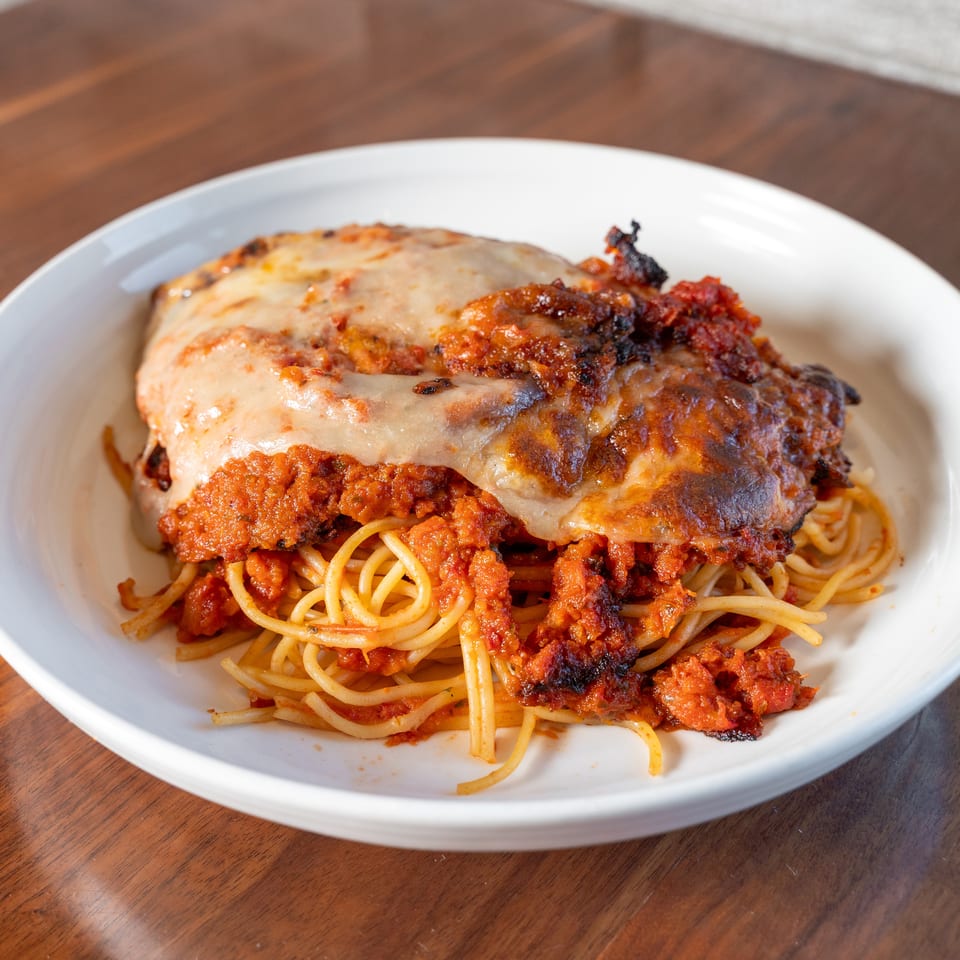 Cheesy breaded chicken parmesan topped with tomato sauce, served over spaghetti on a white plate.