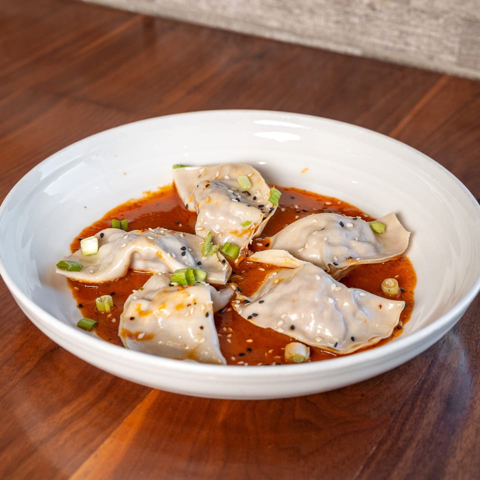 Steamed dumplings in spicy red chili oil, topped with sesame seeds and scallions, served in a white bowl on a wooden table.