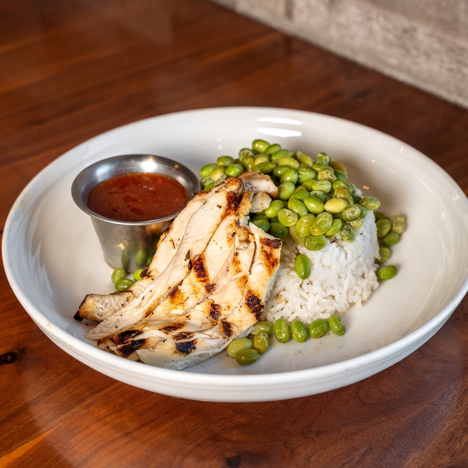 Grilled chicken slices, white rice, green beans, and red dipping sauce served on a white plate.