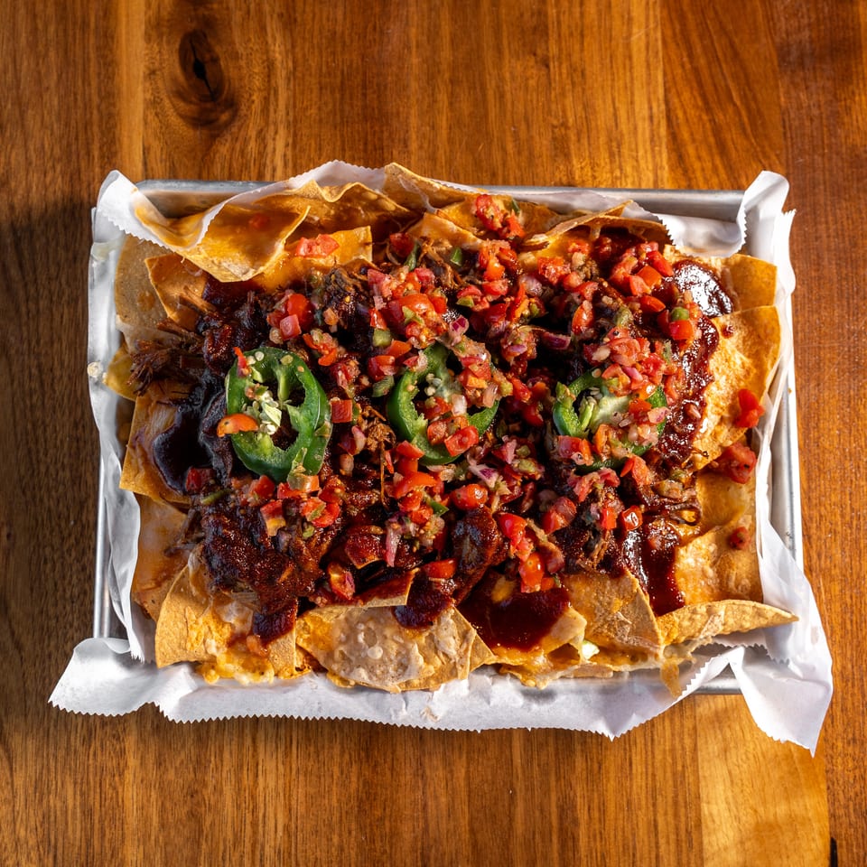 Loaded nachos topped with shredded meat, melted cheese, pico de gallo, sliced jalapeños, and barbecue sauce in a metal tray.