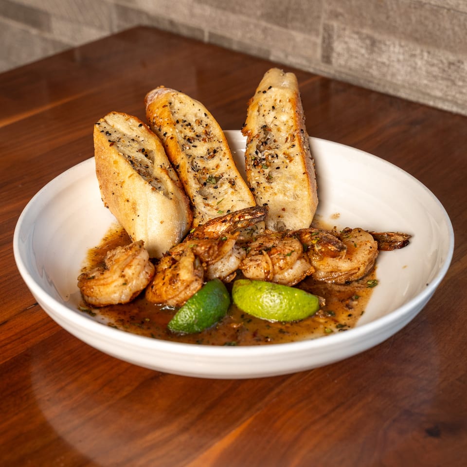 Sautéed shrimp with toasted seasoned bread and lime wedges served in a white bowl on a wooden table.