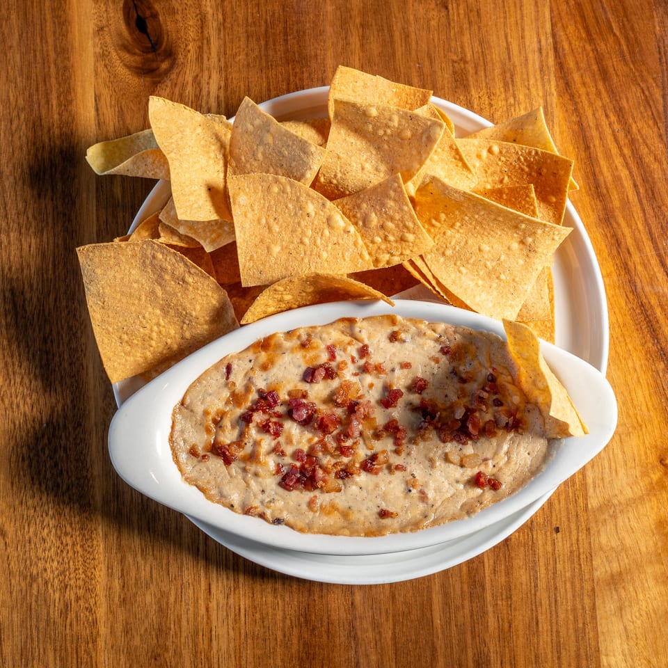 Golden tortilla chips surround a creamy, bacon-topped dip in white serving dishes on a wooden table.