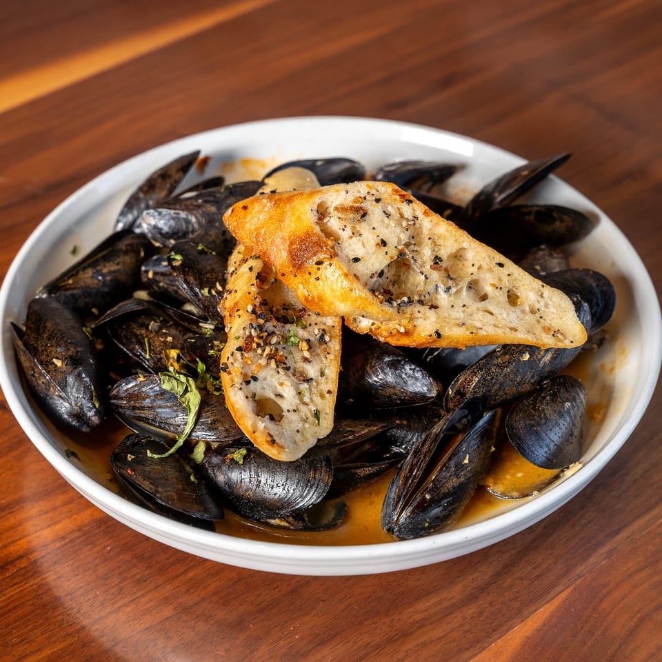 Bowl of mussels in savory broth topped with toasted seeded bread and herbs on a wooden table.