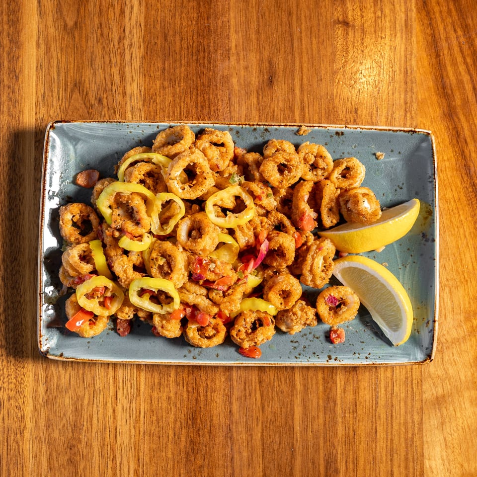 Crispy fried calamari topped with sliced peppers and diced vegetables, served with lemon wedges on a rectangular plate.