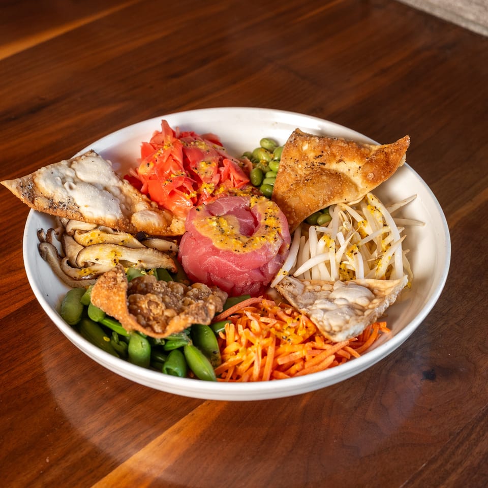 Bowl of colorful Asian-inspired vegetables, mushrooms, shredded carrots, edamame, sliced tuna, and crispy wonton toppings on a wooden table.