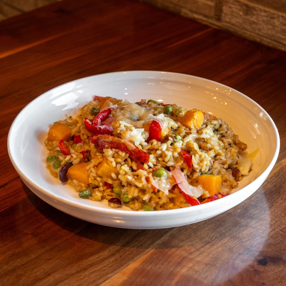 White bowl of savory rice topped with melted cheese, red peppers, peas, squash, and bacon on a wooden restaurant table.