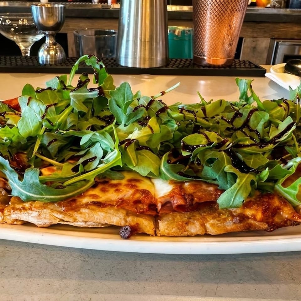 Arugula and prosciutto flatbread pizza with balsamic glaze on a white plate at a bar.