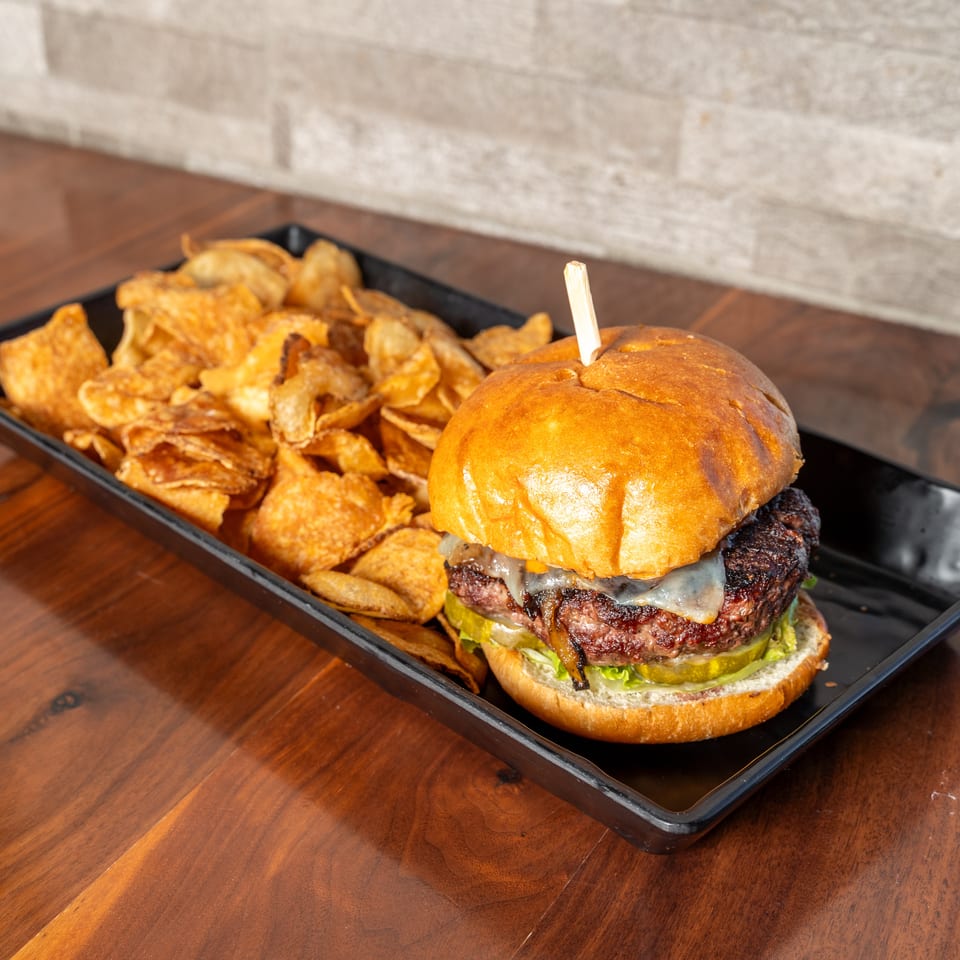 Cheeseburger with lettuce, pickles, and onions served alongside crispy potato chips on a black tray.