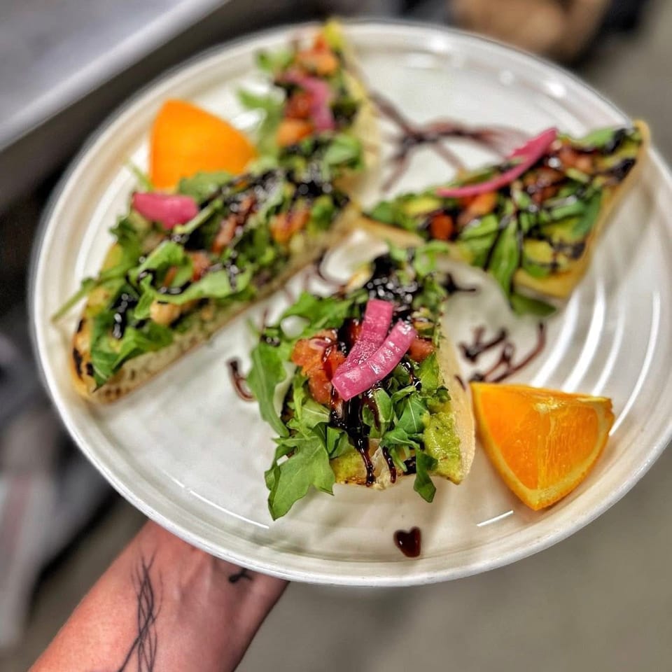 Avocado toast with arugula, tomato, pickled red onion, and balsamic drizzle, served with orange slices.