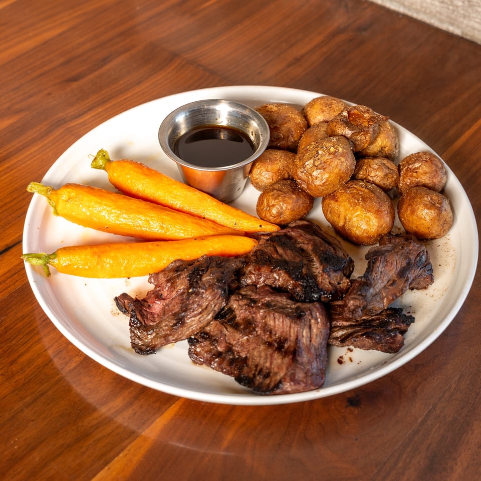 Plate of grilled steak with roasted carrots, baby potatoes, and a small cup of dark dipping sauce on a wooden table.
