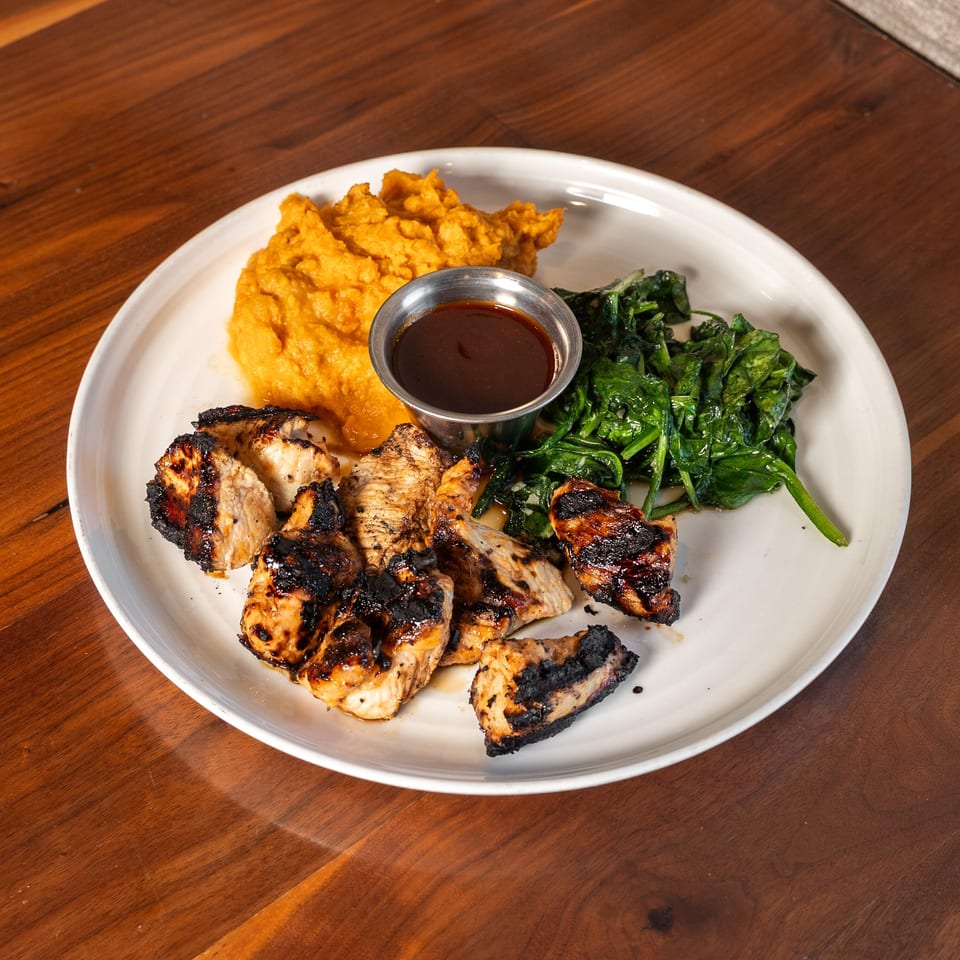 Plate of grilled chicken with mashed sweet potatoes, sautéed greens, and dipping sauce on a wooden table.