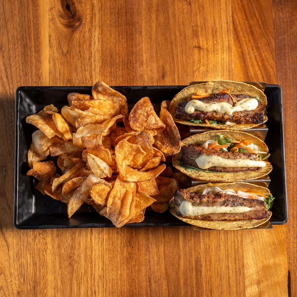 Three grilled meat tacos with creamy sauce, vegetables, and crispy potato chips served on a black platter.