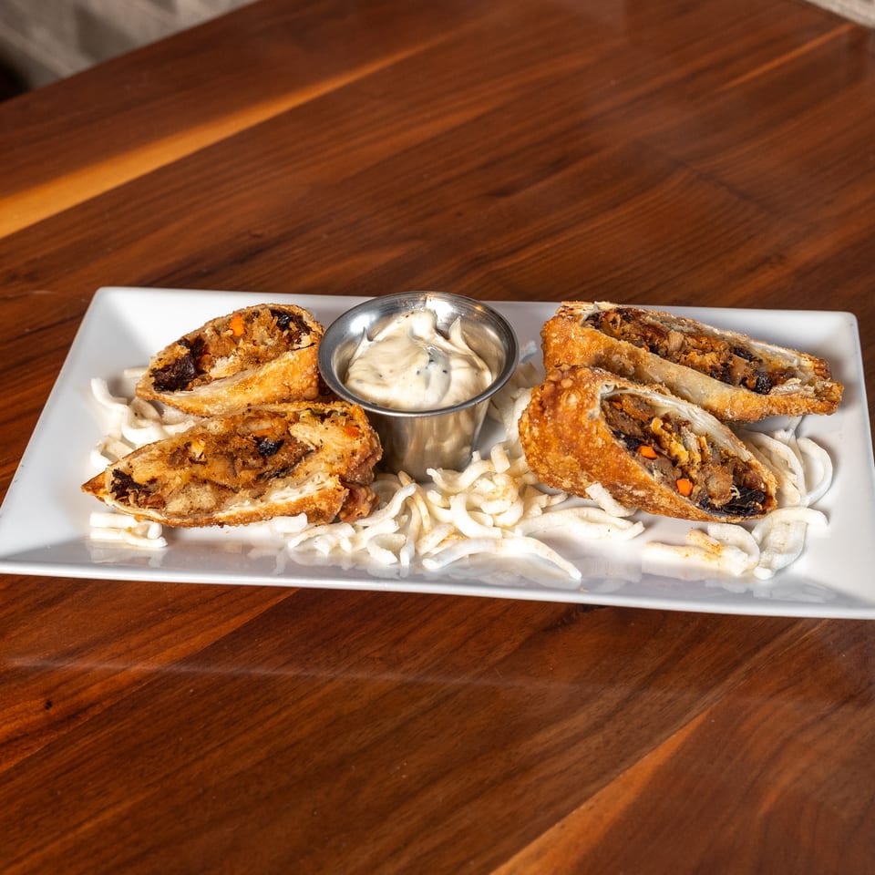 Crispy stuffed pastries served with creamy dipping sauce on a bed of fried noodles atop a white rectangular plate.