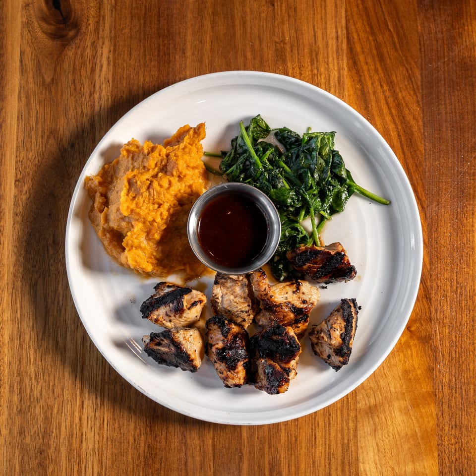 Plate of grilled chicken, mashed sweet potatoes, sautéed greens, and dipping sauce on a wooden table.